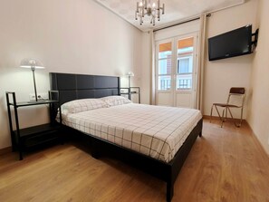Un dormitorio con cama, mesitas de noche, lámpara, silla, televisor y ventana con persianas.