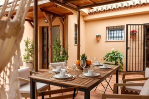 Un área de comedor al aire libre con una mesa de madera preparada para dos personas, rodeada de sillas, plantas en macetas y un muro de azulejos al fondo.