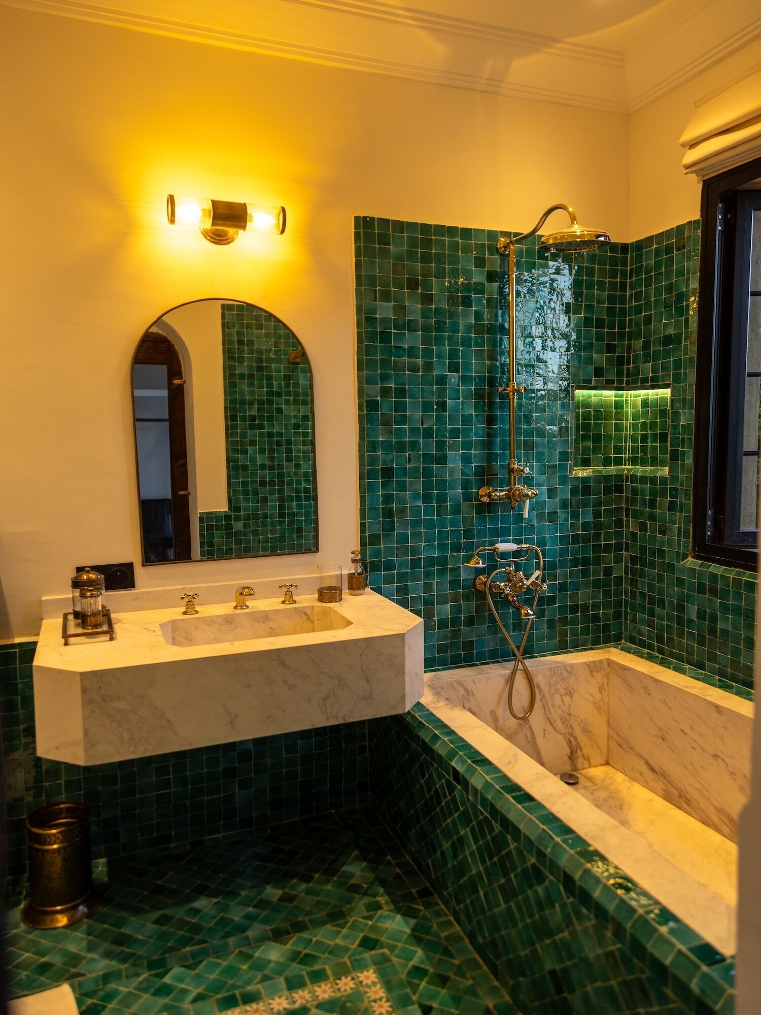 A bathroom with a marble sink, a shower with a golden showerhead, and green mosaic tiles on the walls.