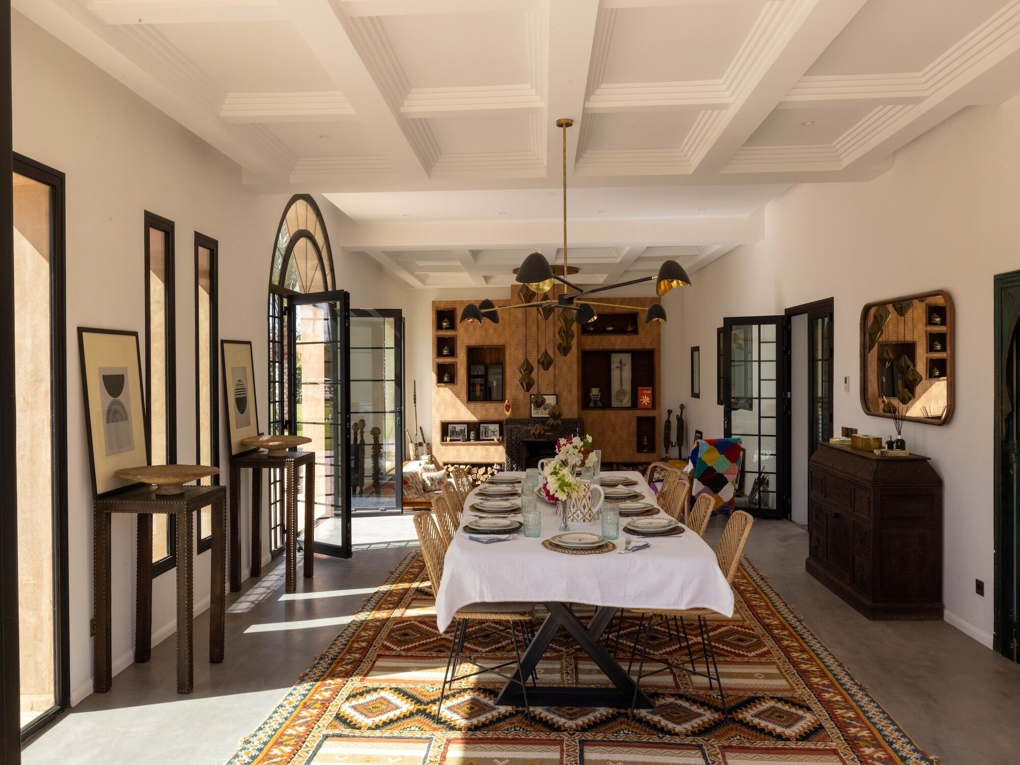 Une salle à manger avec une grande table dressée pour six personnes, entourée de chaises, agrémentée d’un tapis à motifs, d’un lustre et d’œuvres d’art encadrées accrochées aux murs.