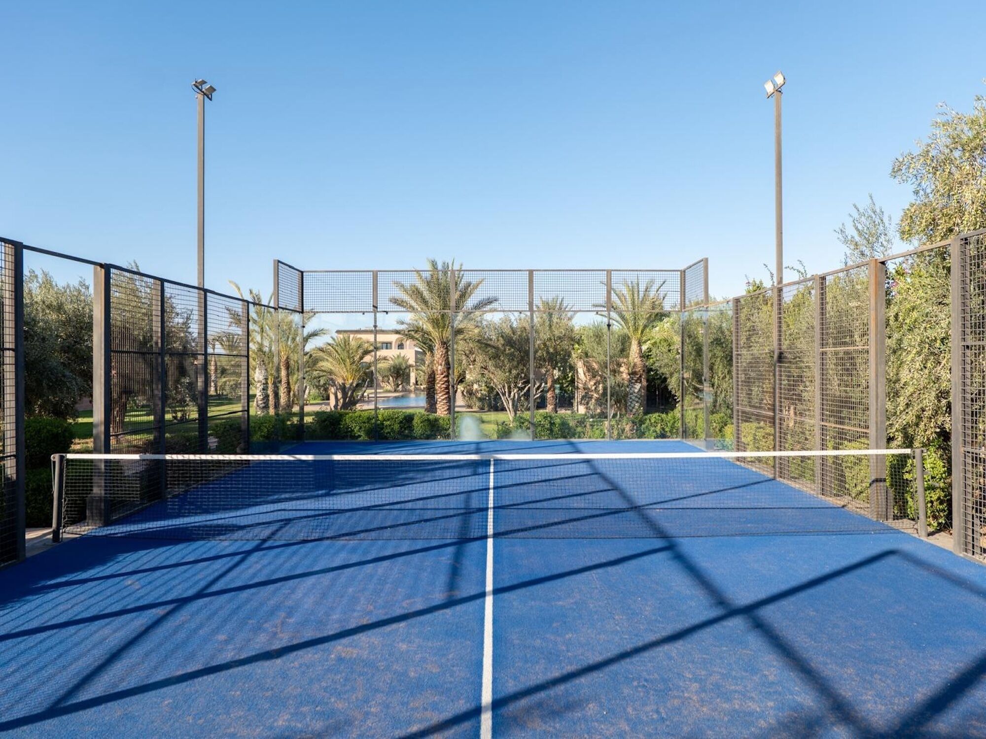 An outdoor tennis court with a net, surrounded by a fence, and palm trees in the background.