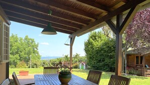 A covered outdoor dining area with a table and chairs, a hanging light fixture, and a view of a garden and distant landscape. - Casale del Sole in the hills of Trevignano: pool and nature 5 min. from the Lake! (Monterosi)