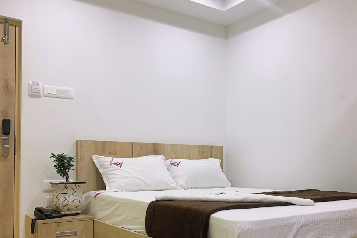 A bedroom with a wooden bed, a nightstand with a phone, a wall-mounted thermostat, and a door with a handle.