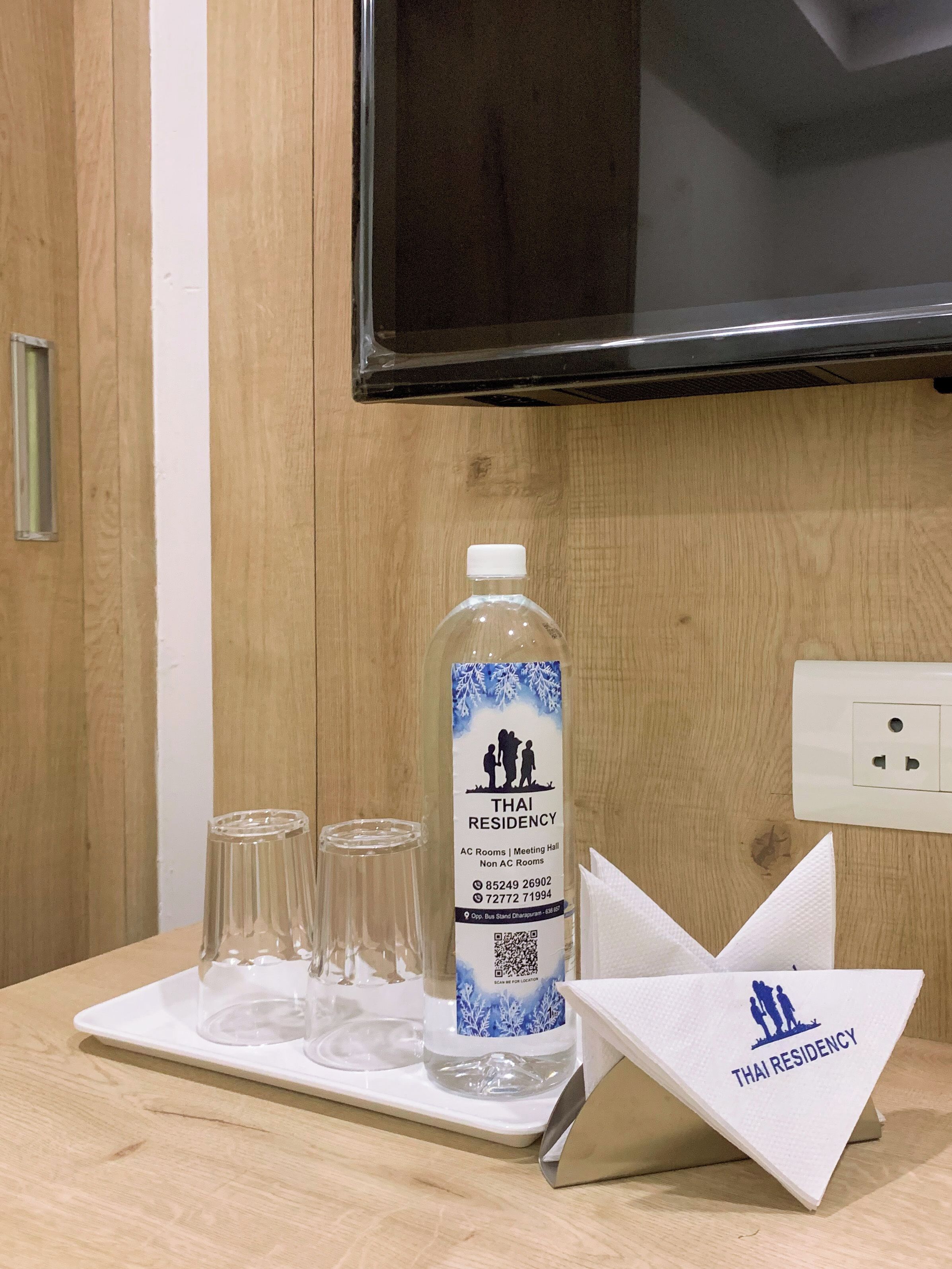Une chambre d’hôtel avec un meuble en bois, un téléviseur fixé au mur, une bouteille d’eau avec une étiquette, et un plateau avec des verres et un porte-serviettes.