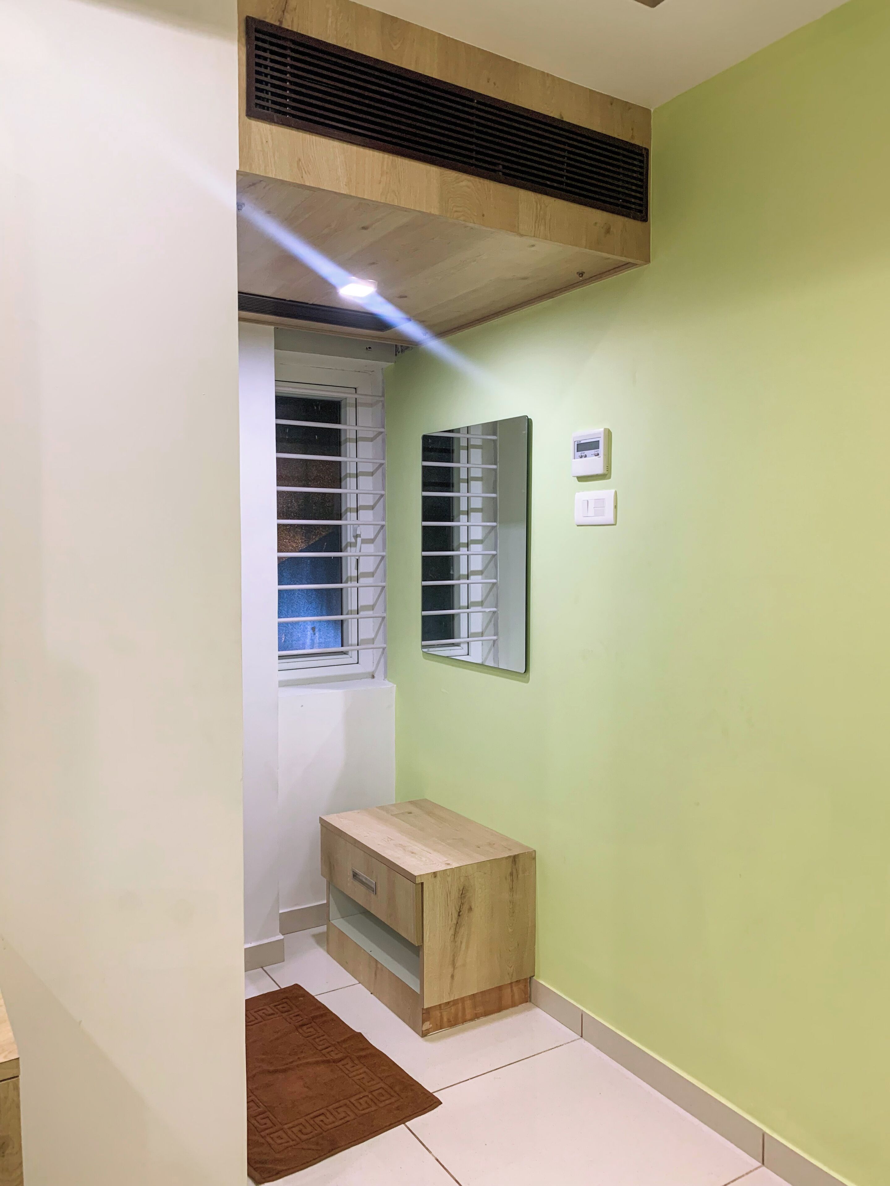 A room with a wooden cabinet, a window with blinds, a wall-mounted thermostat, and a brown rug on the floor.
