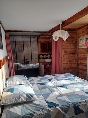 Een slaapkamer met een bed, een houten garderobe, een raam met gordijnen en een klein plankje met decoratieve objecten.