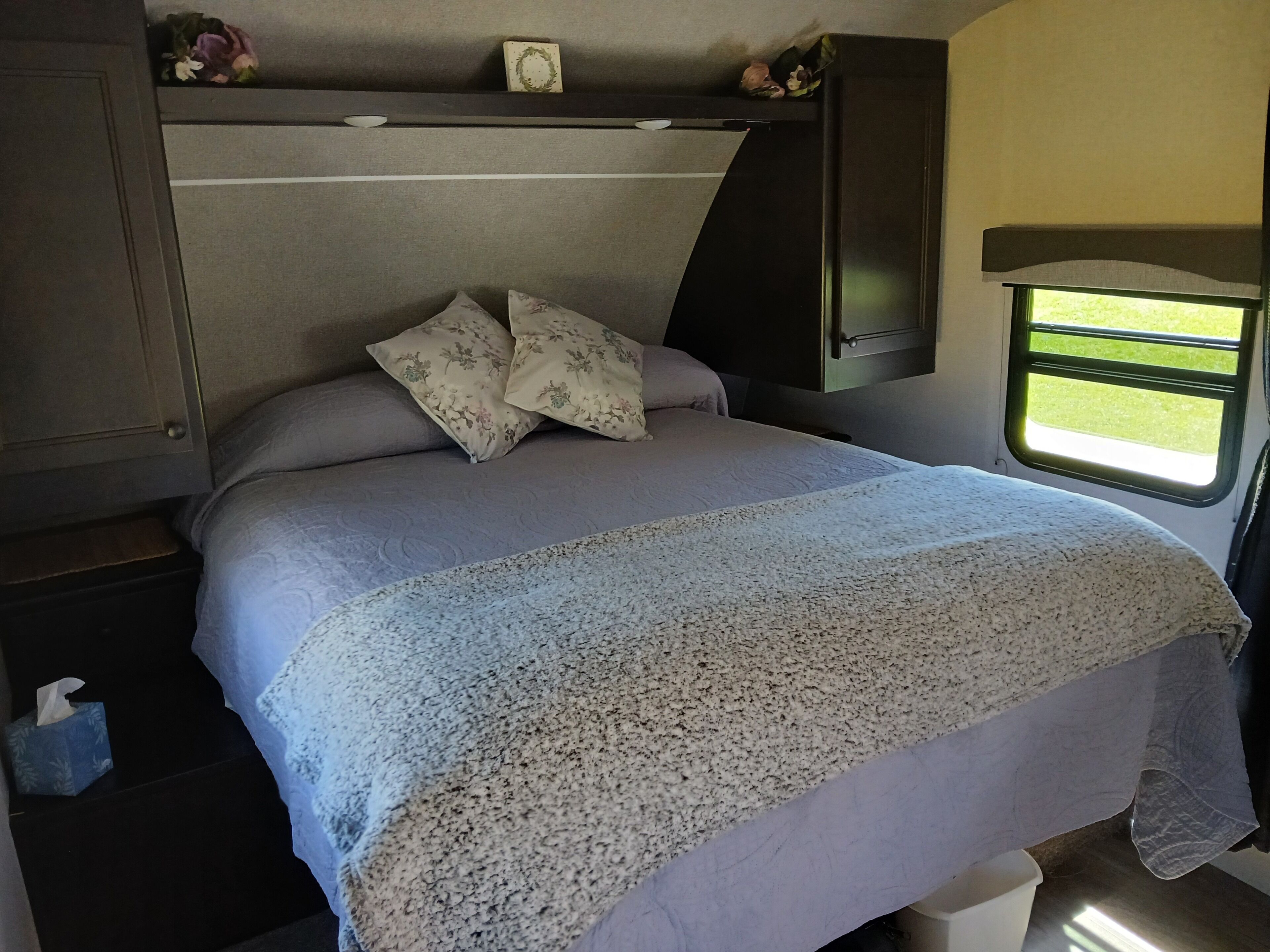 A bed with a floral pillow, a grey headboard, and a window with a view of grass.