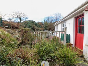 A white building with a red door, surrounded by overgrown vegetation and a wooden fence. - Herons Cove Cottage (Ballydehob)