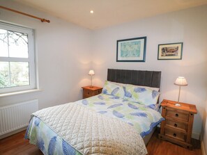 A bedroom with a bed, two bedside tables, a window, and two framed pictures on the wall. - Herons Cove Cottage (Ballydehob)