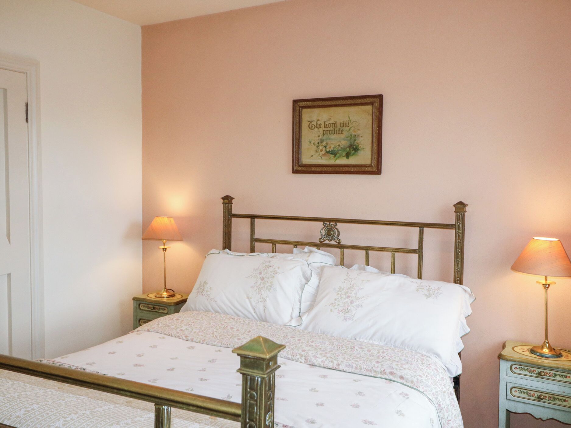 Una cama con una colcha floral, dos lámparas de noche, una cita enmarcada en la pared y una puerta a la izquierda.
