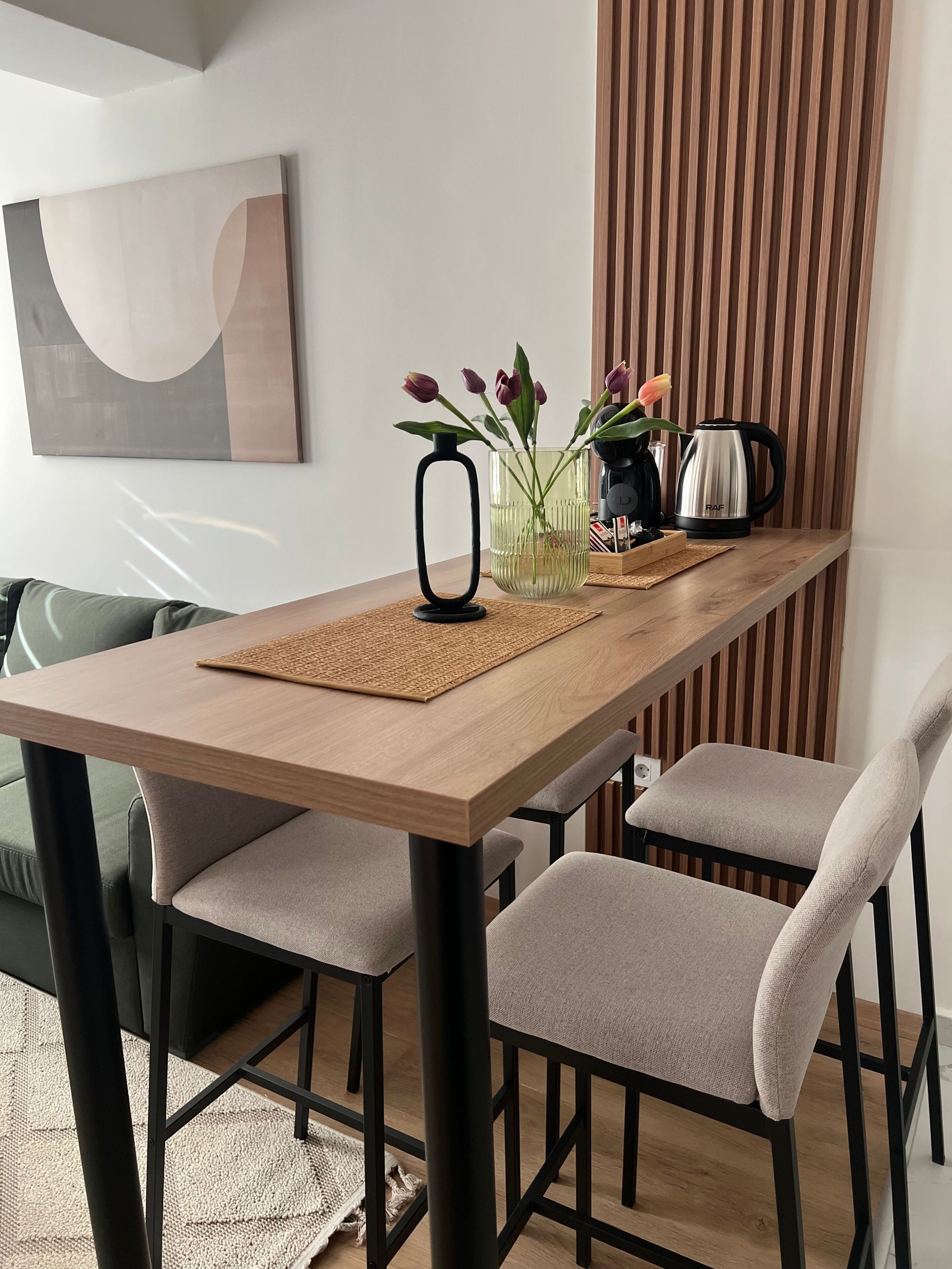 A dining area with a wooden table, four grey chairs, a black vase with flowers, a kettle, and a wall-mounted abstract painting.
