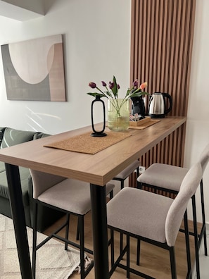 Un coin repas avec une table en bois, quatre chaises grises, un vase noir avec des fleurs, une bouilloire et un tableau abstrait accroché au mur.