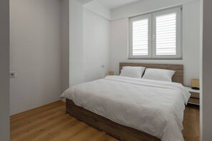 A bedroom with a wooden bed, white bedding, a nightstand, and a window with blinds. - Centrally Located Cozy D&O Apartment (Skopje)