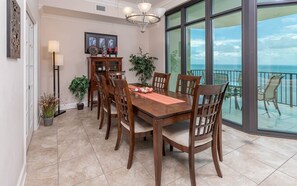 A dining area with a large table, chairs, a chandelier, a cabinet, and a view of the ocean through large windows. - Phoenix West 2605 3 Bedroom Condo (Orange Beach)
