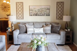 Un salon avec un canapé gris, des coussins décoratifs, un vase de fleurs et des œuvres d’art murales.