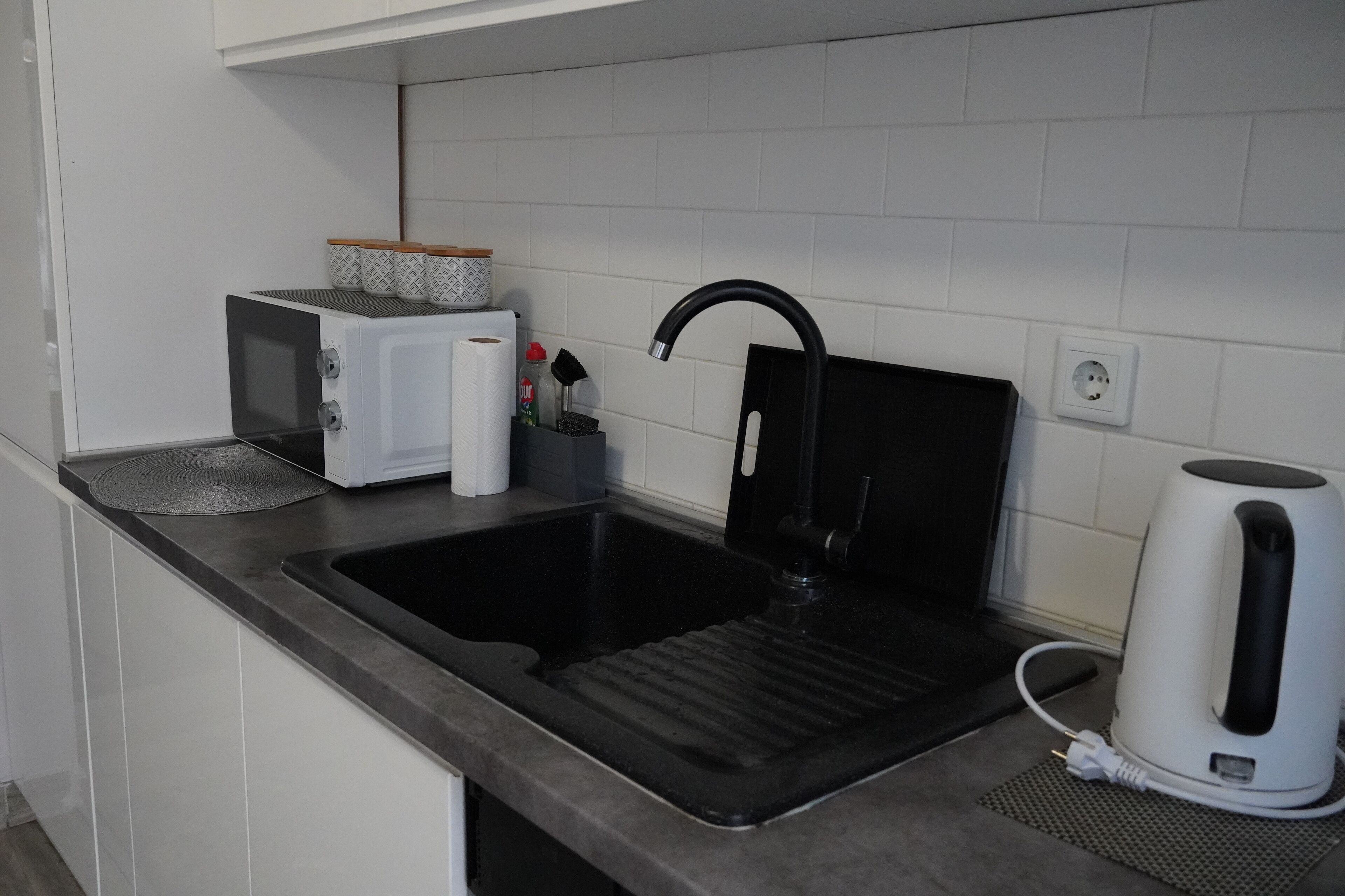 A kitchen with a black sink, a white kettle, a microwave, and a coffee maker on the counter.