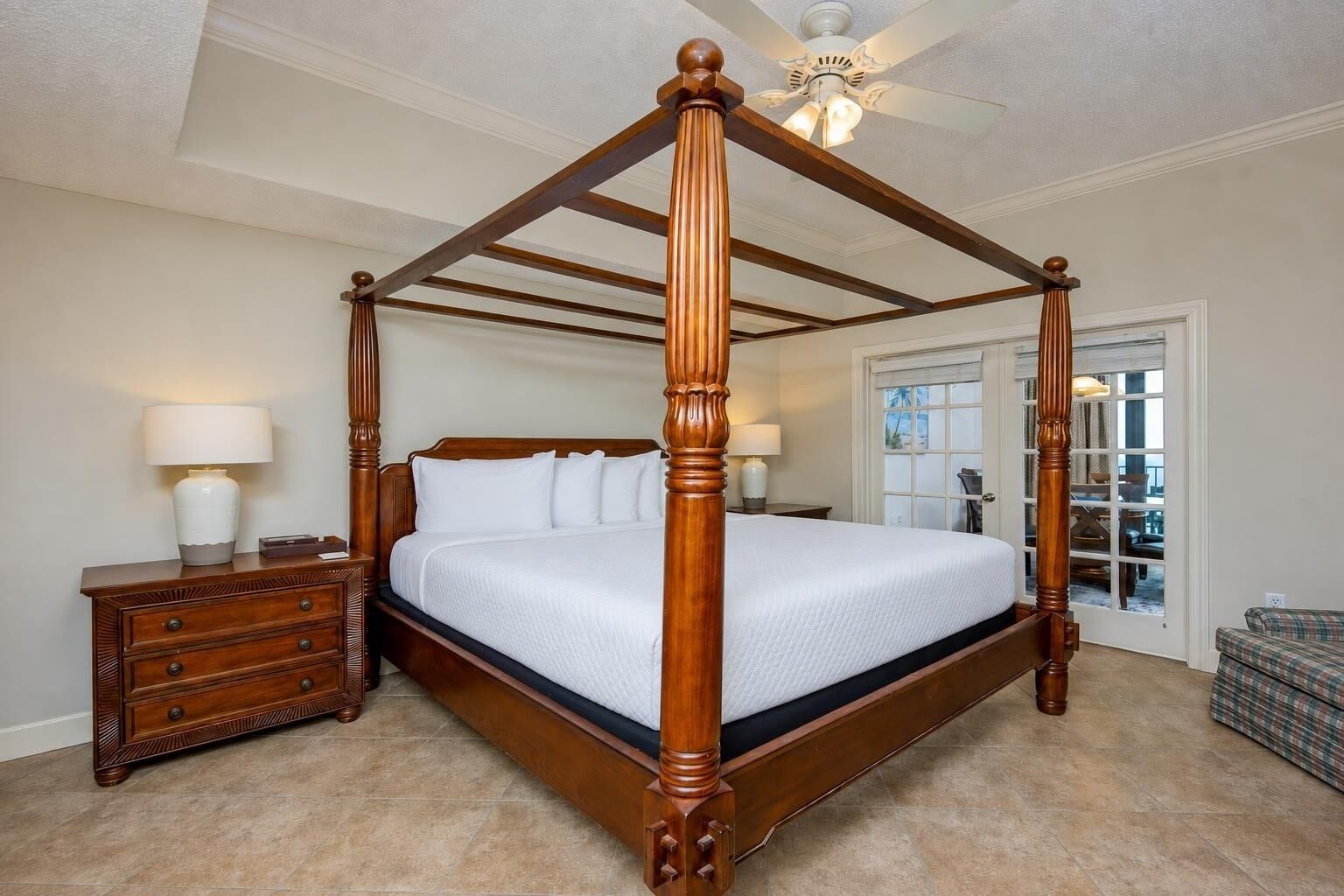 A four-poster bed with a wooden frame, a nightstand with a lamp, and a view of the outside through a door.
