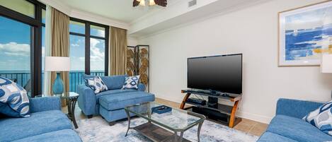 Una sala de estar con un televisor de pantalla plana grande, sofás azules, una mesa de centro de vidrio y un ventilador de techo.