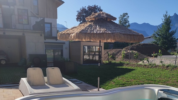 Un jacuzzi con chorros, dos sillones reclinables, una estructura de techo de paja y una casa al fondo.