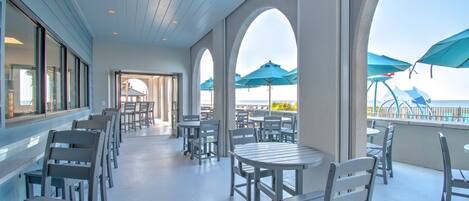 A covered outdoor seating area with tables and chairs, large windows, and blue umbrellas.