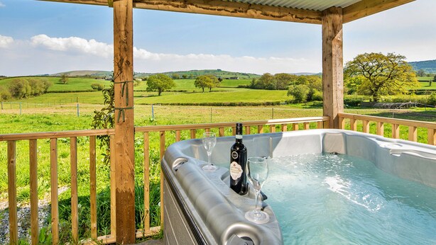 A hot tub with a bottle and glasses, surrounded by a wooden deck and a view of green fields. - Y Golchdy (Llangadog)