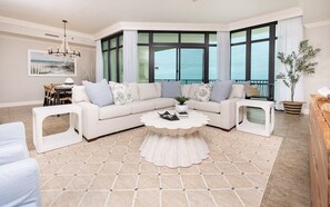 A modern living room with a sectional sofa, a round coffee table, and large windows offering a view. - Phoenix West 2008 3 Bedroom Condo (Orange Beach)