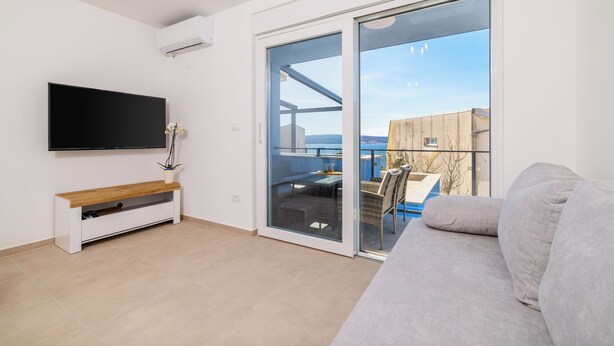 A modern living room with a flat-screen TV, a bench, a sofa, and a sliding glass door leading to an outdoor area with a table and chairs. - Moderne Ferienwohnung mit Meerblick, überdachter Terrasse & Whirlpool (Dramalj)