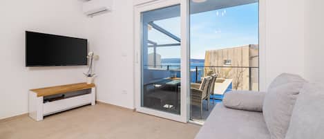 A modern living room with a flat-screen TV, a bench, a sofa, and a sliding glass door leading to an outdoor area with a table and chairs.