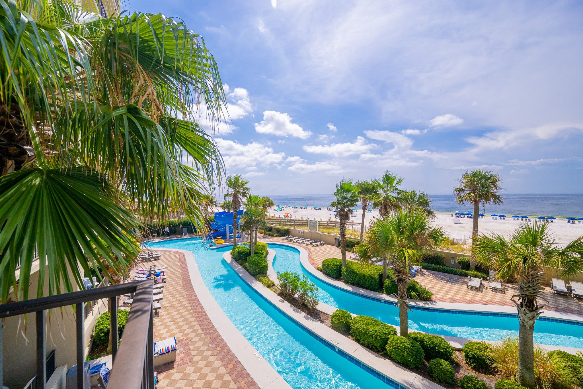 A resort pool area with a view of the beach, lounge chairs, and palm trees.