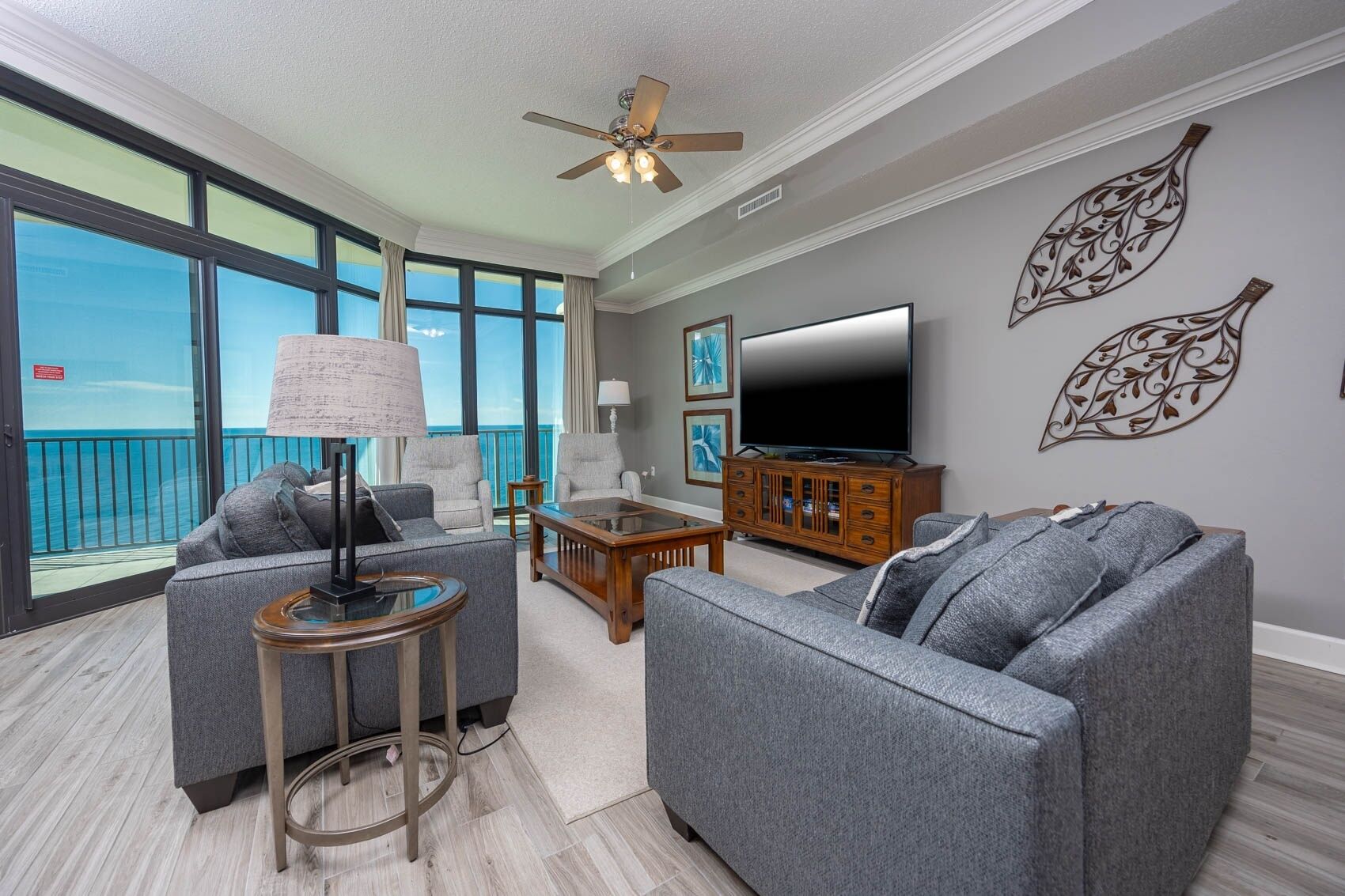 A modern living room with a large TV, grey sofas, a coffee table, and a balcony with a view of the ocean.