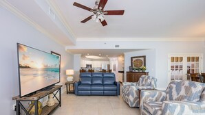 A living room with a large flat-screen TV, a ceiling fan, a sofa, and a patterned armchair. - Phoenix West 2607 3 Bedroom Condo (Orange Beach)