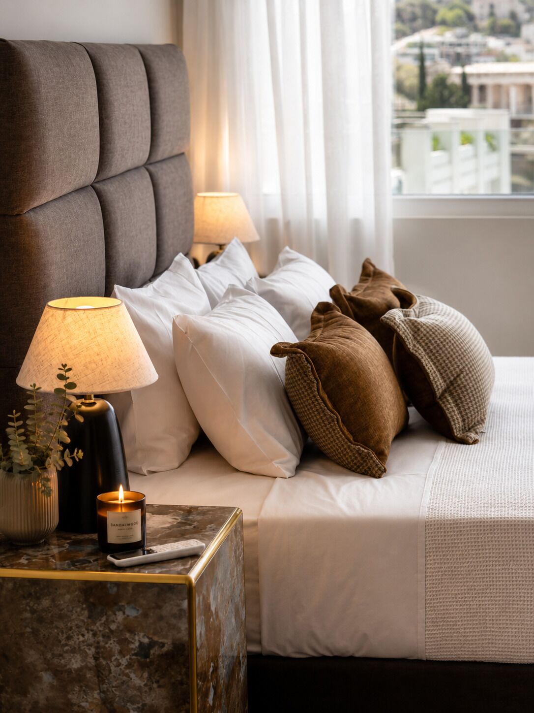 A neatly made bed with white and brown pillows, a bedside lamp, and a candle on a marble nightstand.