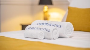 A neatly made bed with a rolled white towel and a pillow with a hotel logo. - Amaris Hotel & Spa by Athens Prime Hotels (Athens)