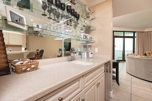 A modern kitchen with a glass display of wine glasses, a built-in blender, and a large mirror. - Phoenix West 1106 3 Bedroom Condo (Orange Beach)