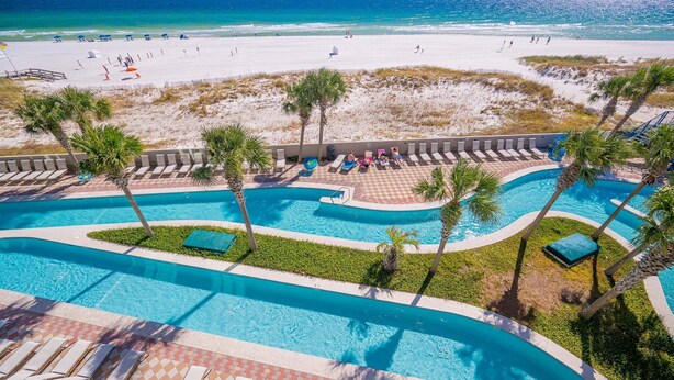 Zona de piscina con varias secciones, palmeras y una playa al fondo.