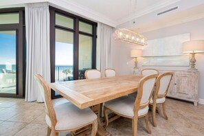 A dining area with a wooden table and chairs, a chandelier, and a view through large windows. - Phoenix West 302 3 Bedroom Condo (Orange Beach)
