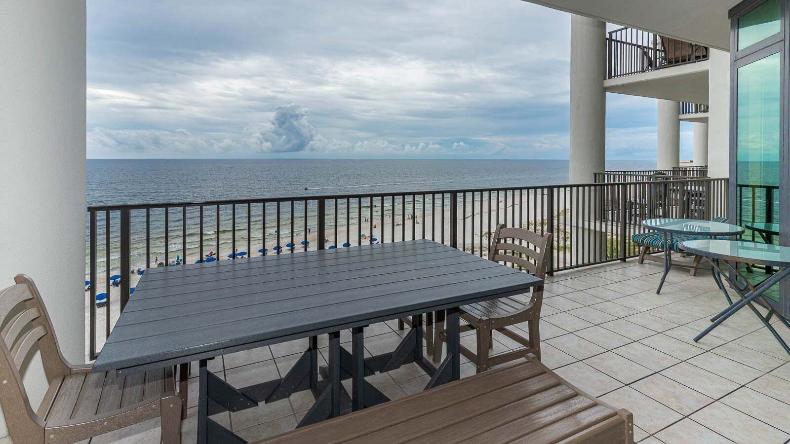 Ein Balkon mit Tisch und Stühlen, der Blick auf einen Strand mit Sonnenschirmen und Menschen freigibt.