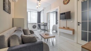 A modern living room with a sofa, a coffee table, a TV mounted on the wall, and a laundry area with a washing machine and dryer.
