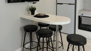 A modern kitchen with a flat-screen TV, a white countertop, four black stools, a refrigerator, and a small oven.