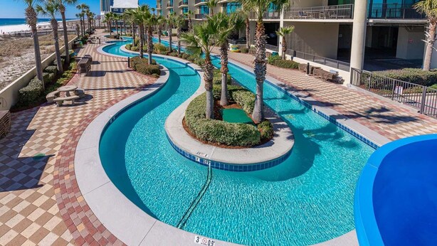 Un área de piscina con diseño curvo, palmeras y vista a un edificio de hotel.