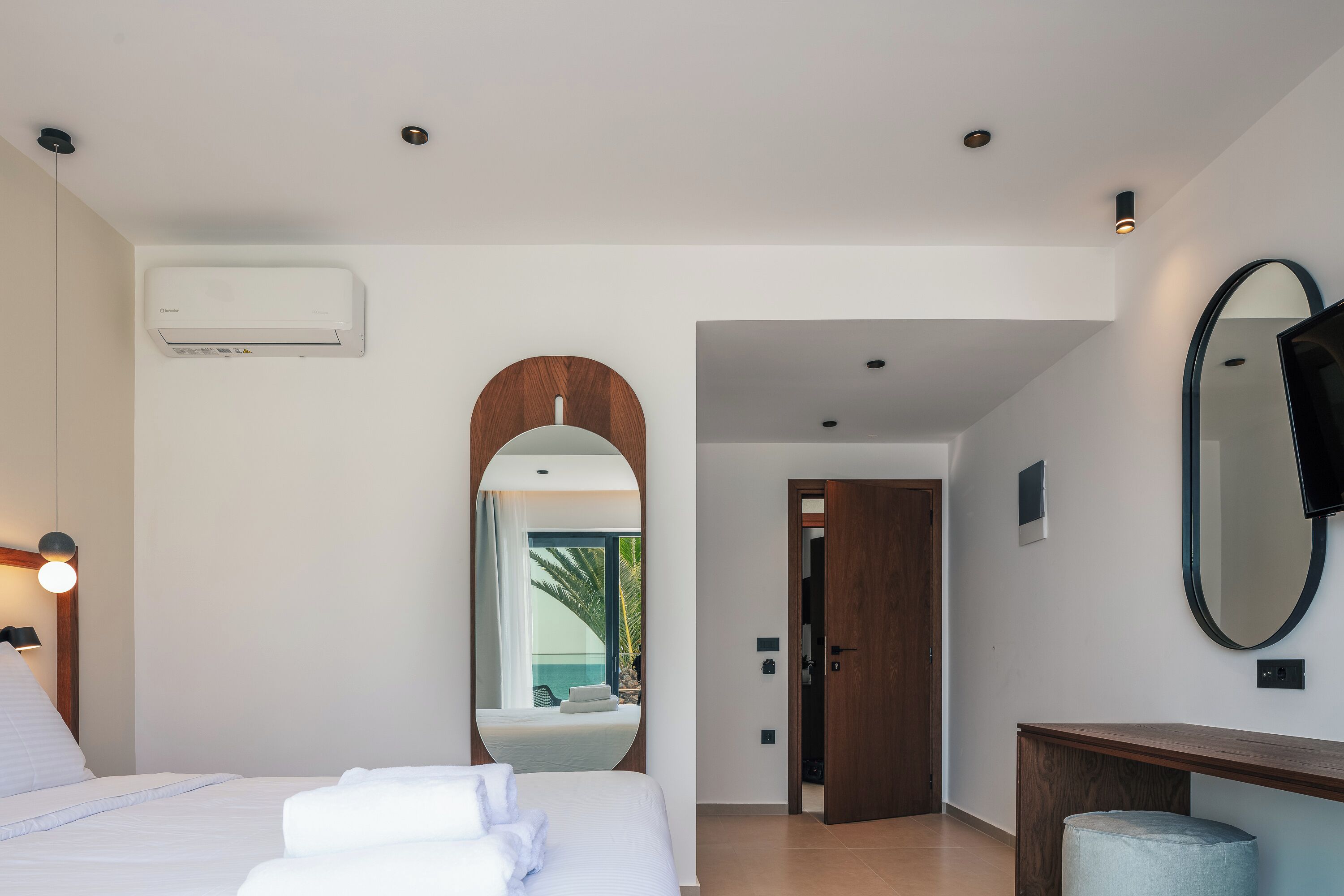 A modern hotel room with a bed, a large mirror, a wooden console table, and a view through an arched doorway.