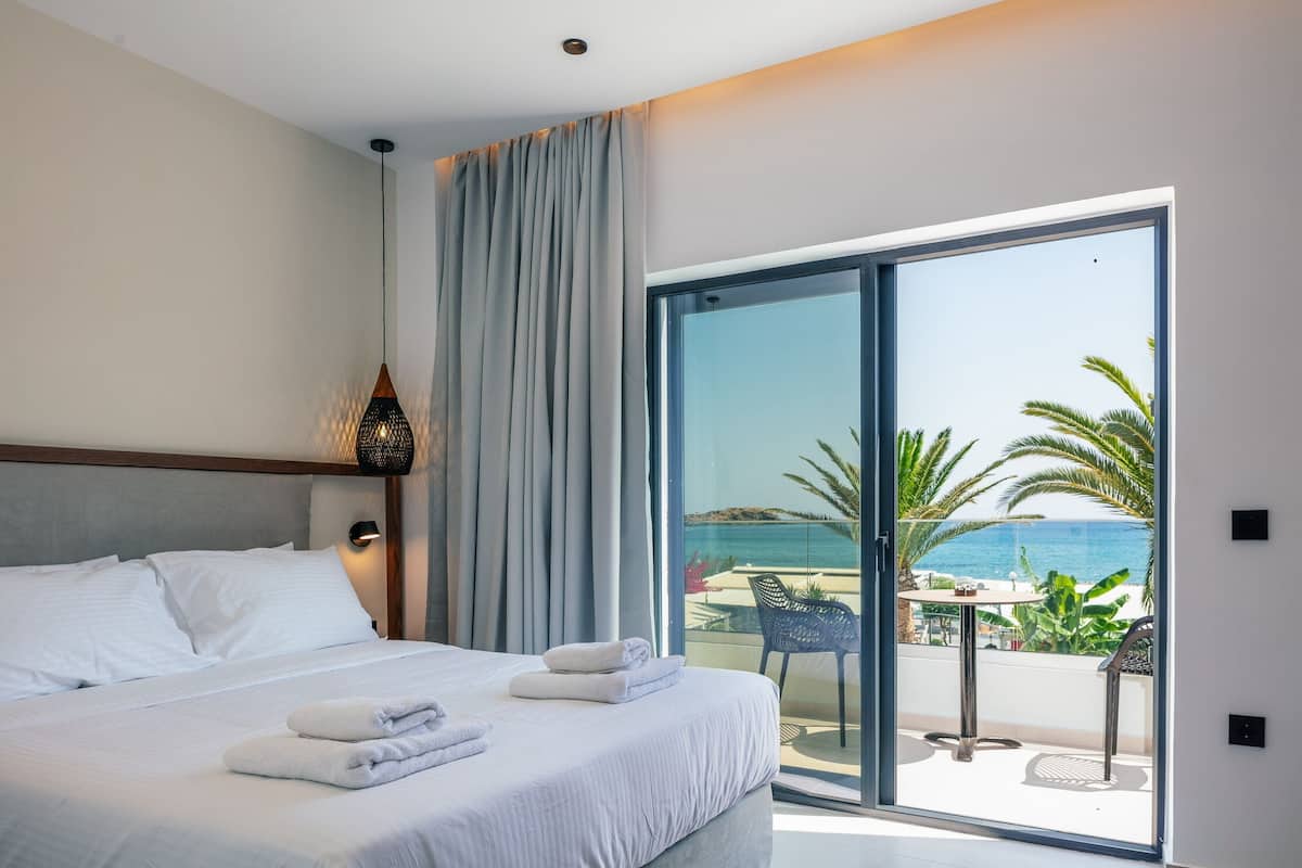 A hotel room with a bed, a view of the sea, and a balcony with outdoor furniture.