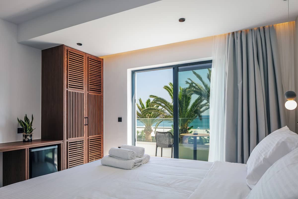 A modern hotel room with a bed, a small desk, a wardrobe, and a balcony with a view of palm trees.