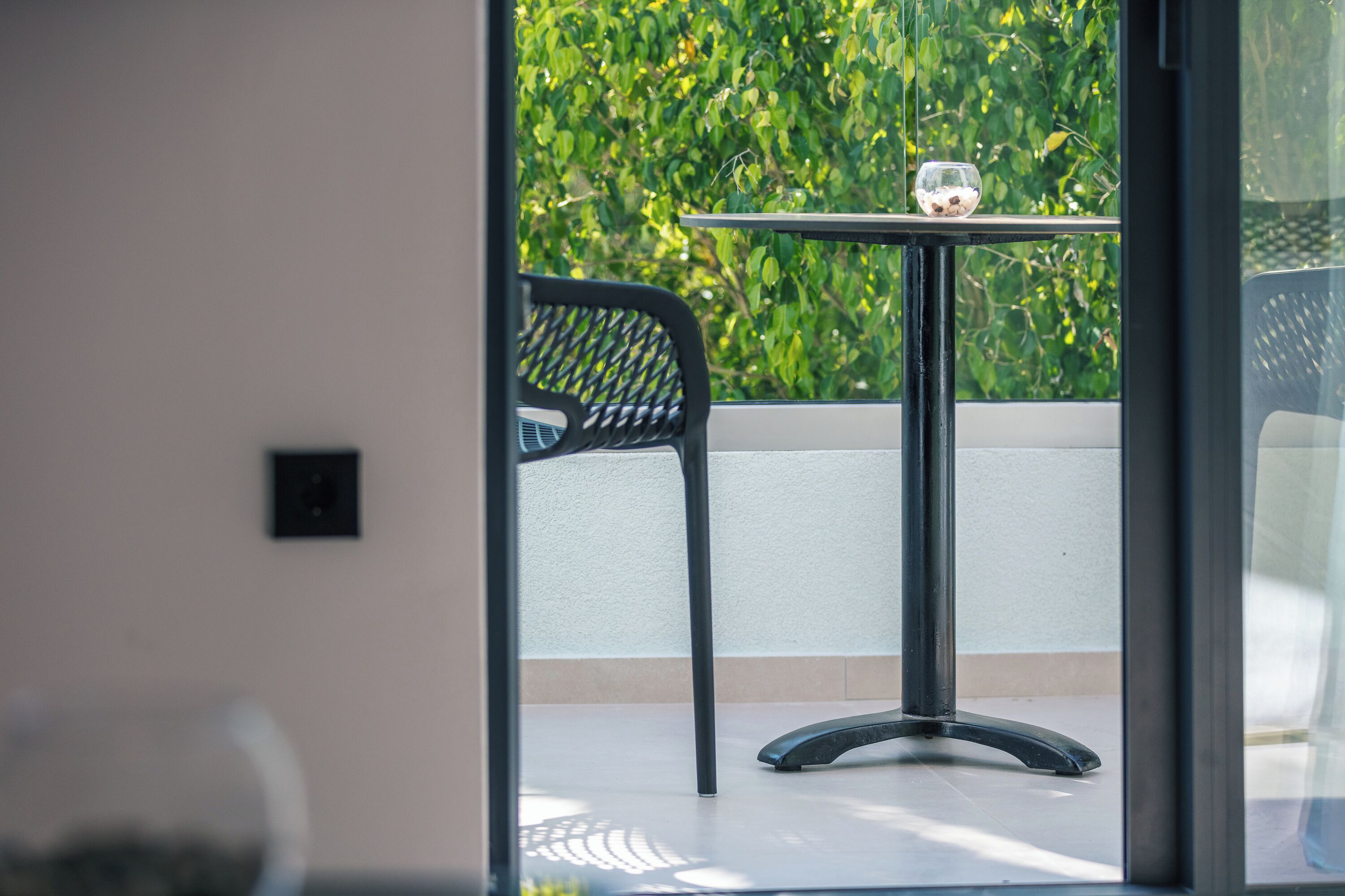 A view of a balcony with a table and chair, a small glass bowl on the table, and a green hedge in the background.