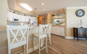 A kitchen with white cabinetry, a bar area with white chairs, and a dining table. - Phoenix West 2507 3 Bedroom Condo (Orange Beach)