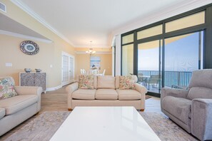 A living room with a beige sofa, a white coffee table, and a gray armchair. There is a large window with a view of the outdoors. - Phoenix West 2507 3 Bedroom Condo (Orange Beach)
