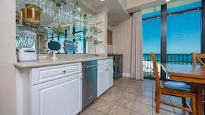 A kitchen with a dishwasher, a countertop with glassware, and a dining area with a table and chairs. - Phoenix West 1609 4 Bedroom Condo (Orange Beach)