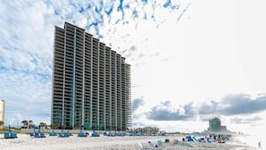 A high-rise hotel with multiple balconies, a beach with sun loungers, and a clear sky. - The Oasis 1815 Formerly Phoenix West Ii 1815 3 Bedroom Condo (Orange Beach)
