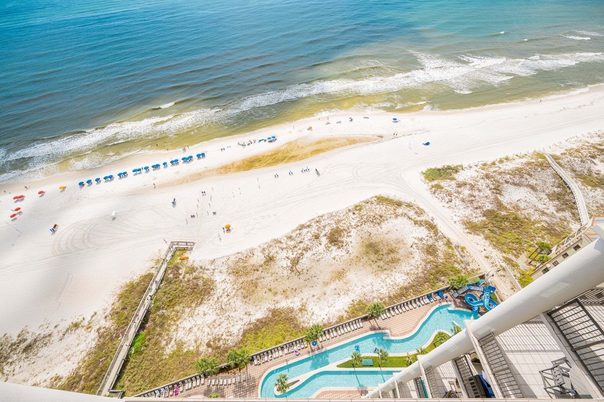An aerial view of a beach with a swimming pool, beach umbrellas, and a boardwalk.