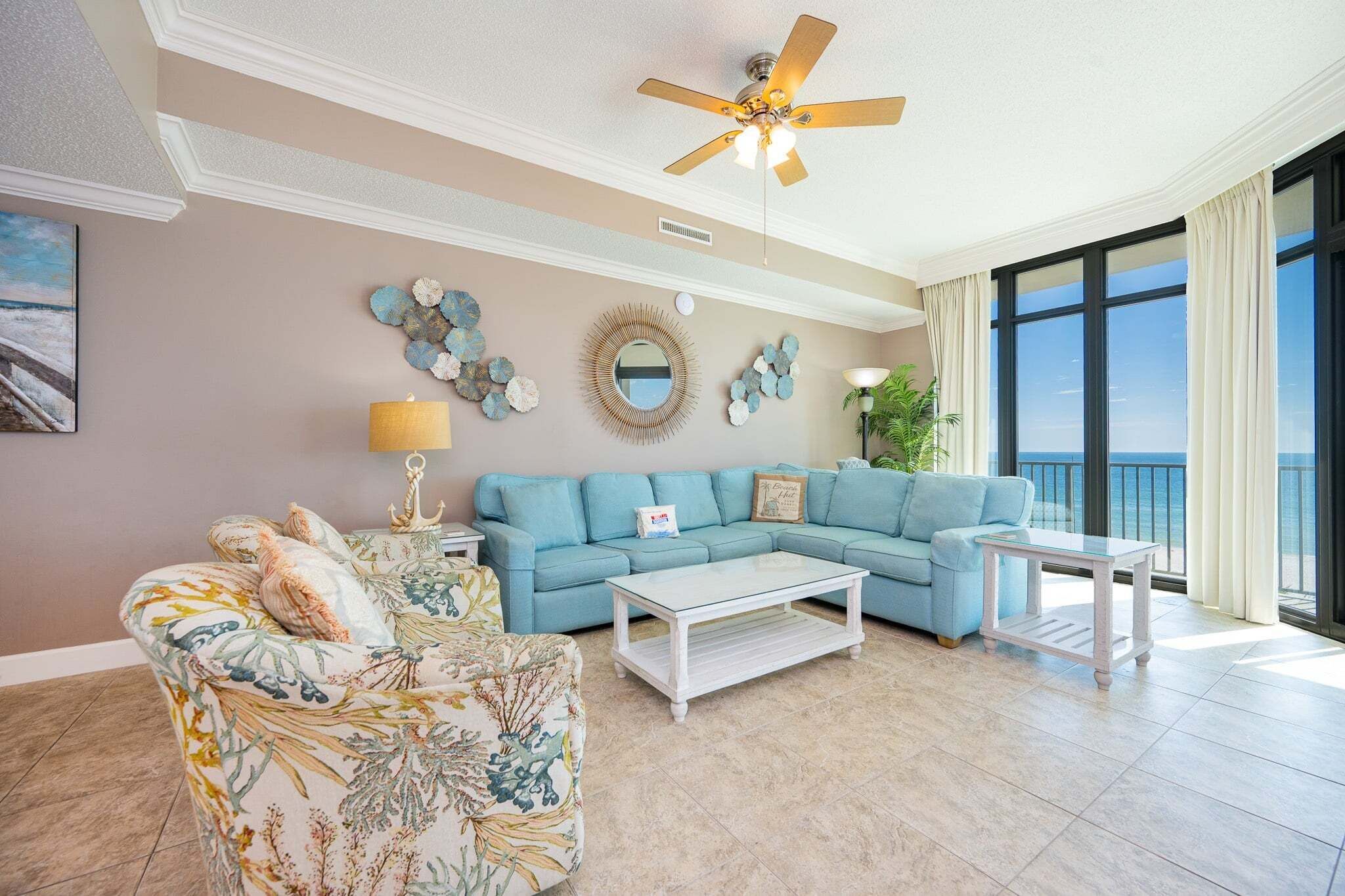 A living room with a blue sofa, a floral armchair, a white coffee table, a ceiling fan, and a large window with curtains.
